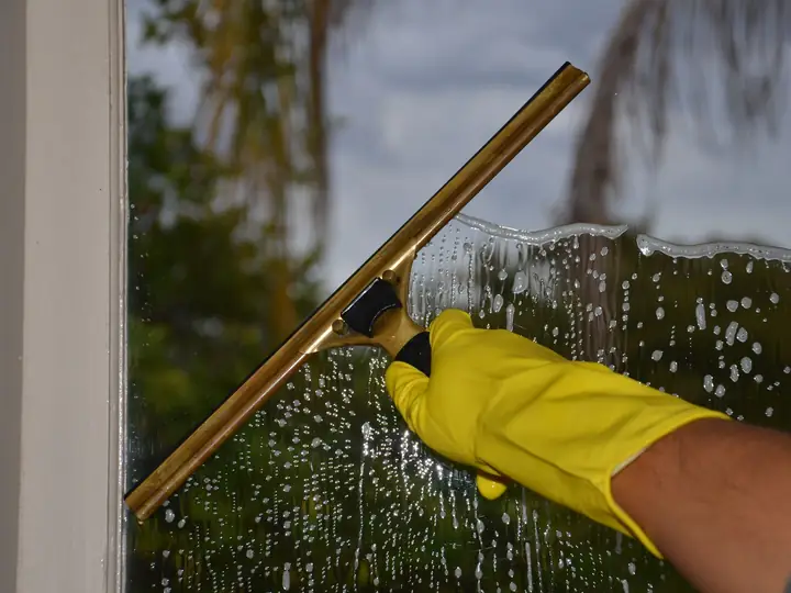 Hand mit gelbem Handschuh zieht mit einem Abzieher das Wasser von einer nassen Fensterscheibe